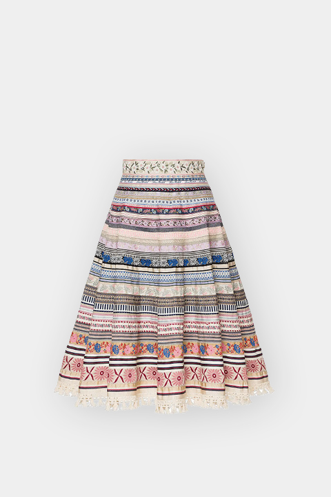 Una gonna al ginocchio a trapezio con strisce orizzontali colorate, motivi intrecciati e ricami floreali su sfondo bianco.