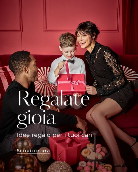 Una donna sorridente, un uomo e un bambino sono seduti insieme in una festosa stanza rossa e scartano un pacco regalo rosa. Diverse caramelle sono su un tavolo nelle vicinanze. Il testo italiano recita: "Regala gioia. Idee regalo per i tuoi cari. Scopri ora.".