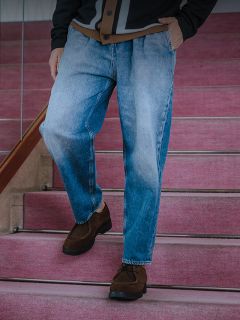 Un uomo con una maglietta bianca, una giacca di jeans scura e jeans è in piedi accanto a una ringhiera appoggiata a un muro di cemento e a una recinzione, sopra una tenda gialla.