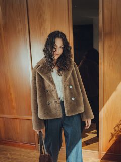 Una donna con i capelli ricci che indossa un cappotto di pelliccia sintetica marrone, una camicetta bianca e jeans a gamba larga, in piedi in un interno con una borsa marrone in mano, con pannelli di legno e luce solare sullo sfondo.