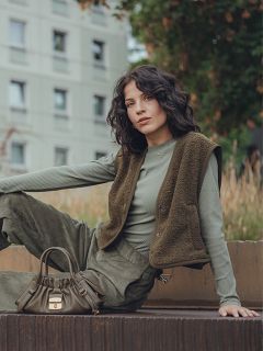 Una donna con capelli scuri ondulati è seduta all'aperto su una superficie rialzata, indossa un top a coste verde a maniche lunghe, un gilet color oliva e pantaloni abbinati. Ha una piccola borsetta color oliva accanto a sé e un edificio residenziale sullo sfondo.