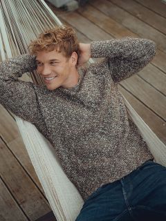 Una persona sorridente con i capelli ricci, che indossa un maglione di lana marrone e jeans blu, è sdraiata su un'amaca bianca su un ponte di legno con le mani dietro la testa.