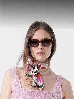 Donna in un abito rosa strutturato con motivi a rosetta, con occhiali da sole scuri e una sciarpa colorata legata al collo, in piedi davanti a uno sfondo semplice.