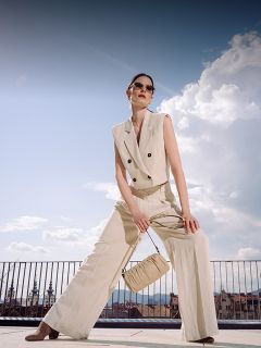 Una donna in un tailleur pantalone beige senza maniche e con occhiali da sole posa all'aperto, con una borsa abbinata in mano, con un paesaggio urbano e un cielo blu sullo sfondo.