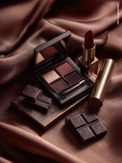 Una palette di ombretti, un rossetto rosso in un astuccio dorato e pezzi di cioccolato fondente sono disposti su un tessuto di seta marrone, creando una composizione lussuosa ed elegante.
