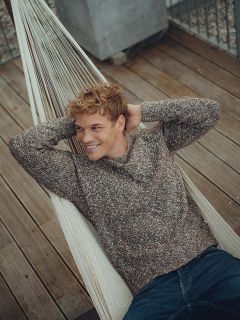 Un giovane uomo con capelli biondi ricci, indosso un maglione marrone e jeans, si rilassa su un'amaca, sorride e guarda di lato. L'amaca è su un ponte di legno con una ringhiera di metallo sullo sfondo.