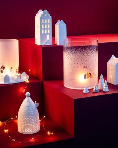 Case di ceramica, alberi e portacandele sono disposti su gradini rossi e sono delicatamente illuminati da catene luminose e luce di candela. La scena ricorda un'accogliente e festosa atmosfera di un villaggio invernale.