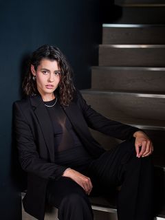 Una donna in un abito nero è seduta su una scala di legno davanti a una parete scura e guarda con sicurezza la fotocamera, i suoi capelli sono acconciati in onde morbide.