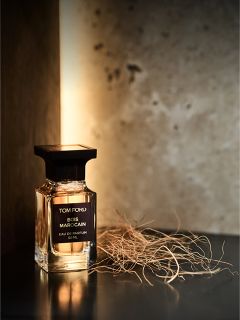 Un flacone del profumo Tom Ford Bois Marocain è posizionato su una superficie scura accanto a erba secca, con uno sfondo caldo e delicatamente illuminato.