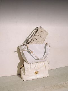 Tre eleganti borse beige e bianche sono impilate ordinatamente davanti a una parete chiara e un pavimento in cemento, illuminate dalla luce naturale del sole.