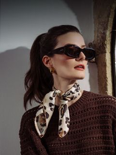 Donna con occhiali da sole scuri, orecchini dorati, una sciarpa con motivo leopardato e un maglione marrone strutturato, che posa vicino a una finestra con la luce sul viso.