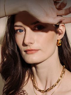 Donna con la pelle chiara e i capelli castani, che indossa gioielli d'oro e tiene la mano sopra gli occhi, uno dei quali è blu e l'altro marrone.