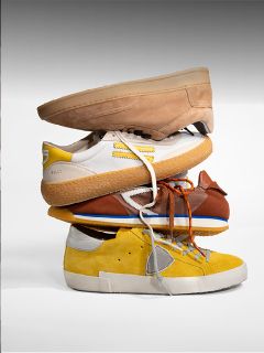 Quattro diverse scarpe da ginnastica nei colori marrone chiaro, bianco, marrone e giallo sono impilate una sopra l'altra davanti a uno sfondo monocolore.