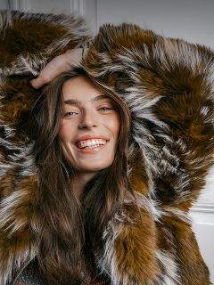 Una donna sorridente che indossa un cappotto di pelliccia sintetica marrone e bianca a pelo lungo, con le mani incrociate dietro la testa.