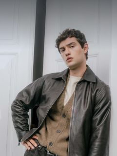 Un uomo che indossa una giacca di pelle scura sopra un cardigan marrone chiaro e una camicia color crema, appoggiato a uno stipite.